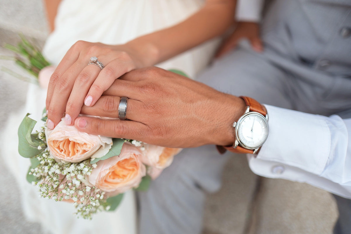 Manos de novios con anillos de matrimonio y ramo de novia de rosas color durazno